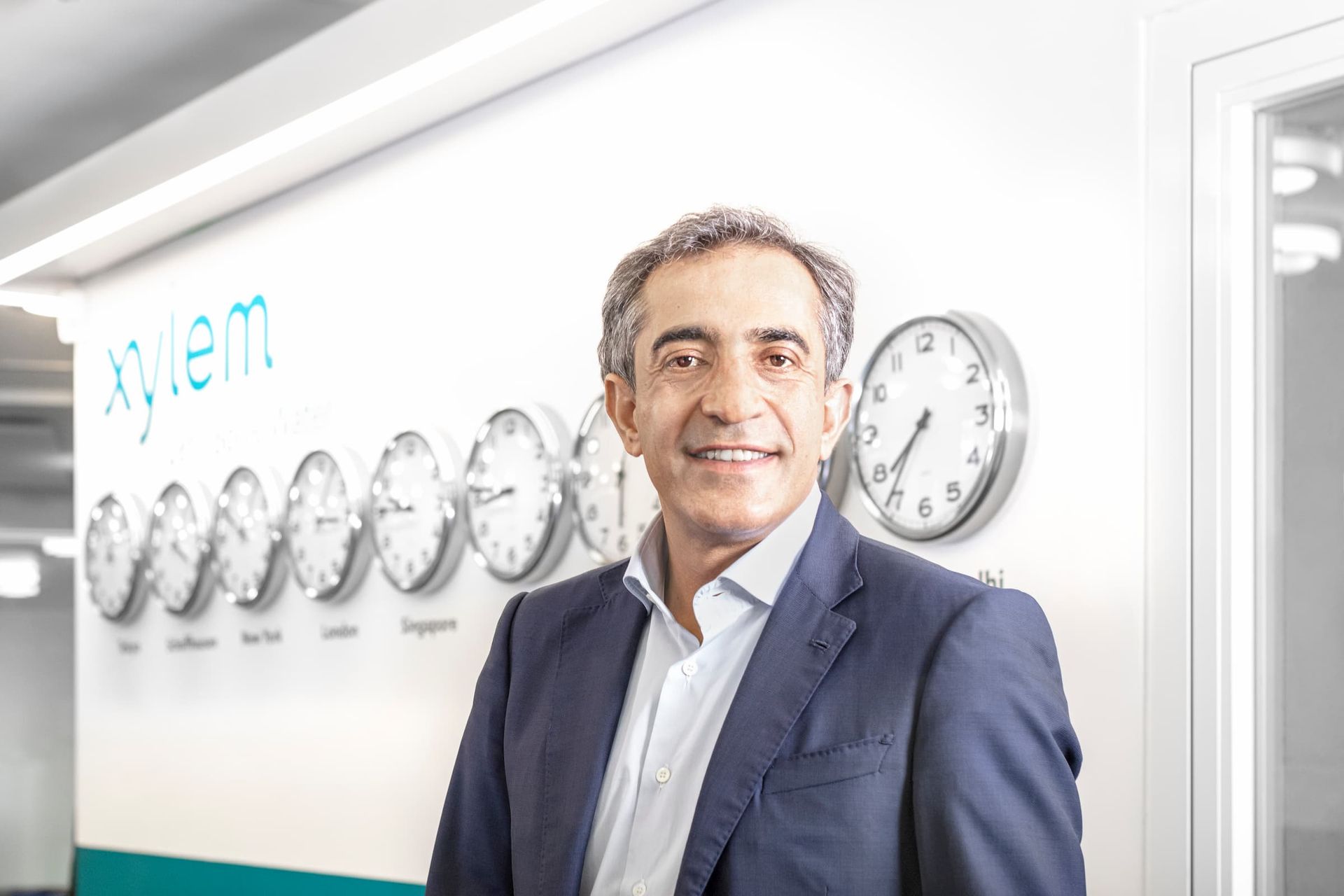 A man in a suit is standing in front of a wall with clocks on it.