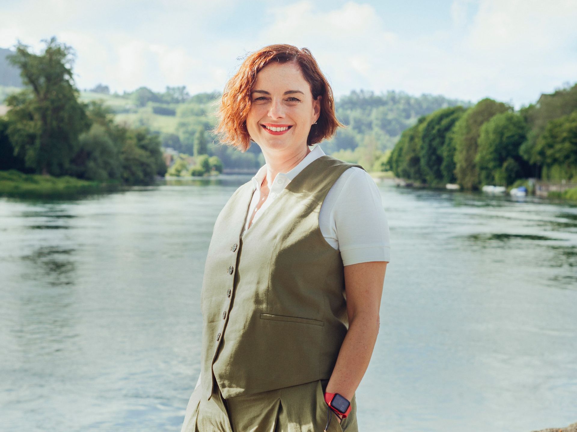 Eine Frau steht mit den Händen in den Taschen neben einem Fluss.