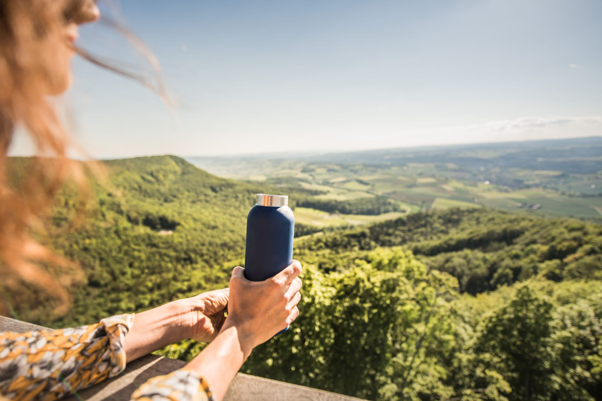 Woman holding a blue water bottle, overlooking a lush green valley in the Randen, Schaffhausen, Switzerland on a sunny day.