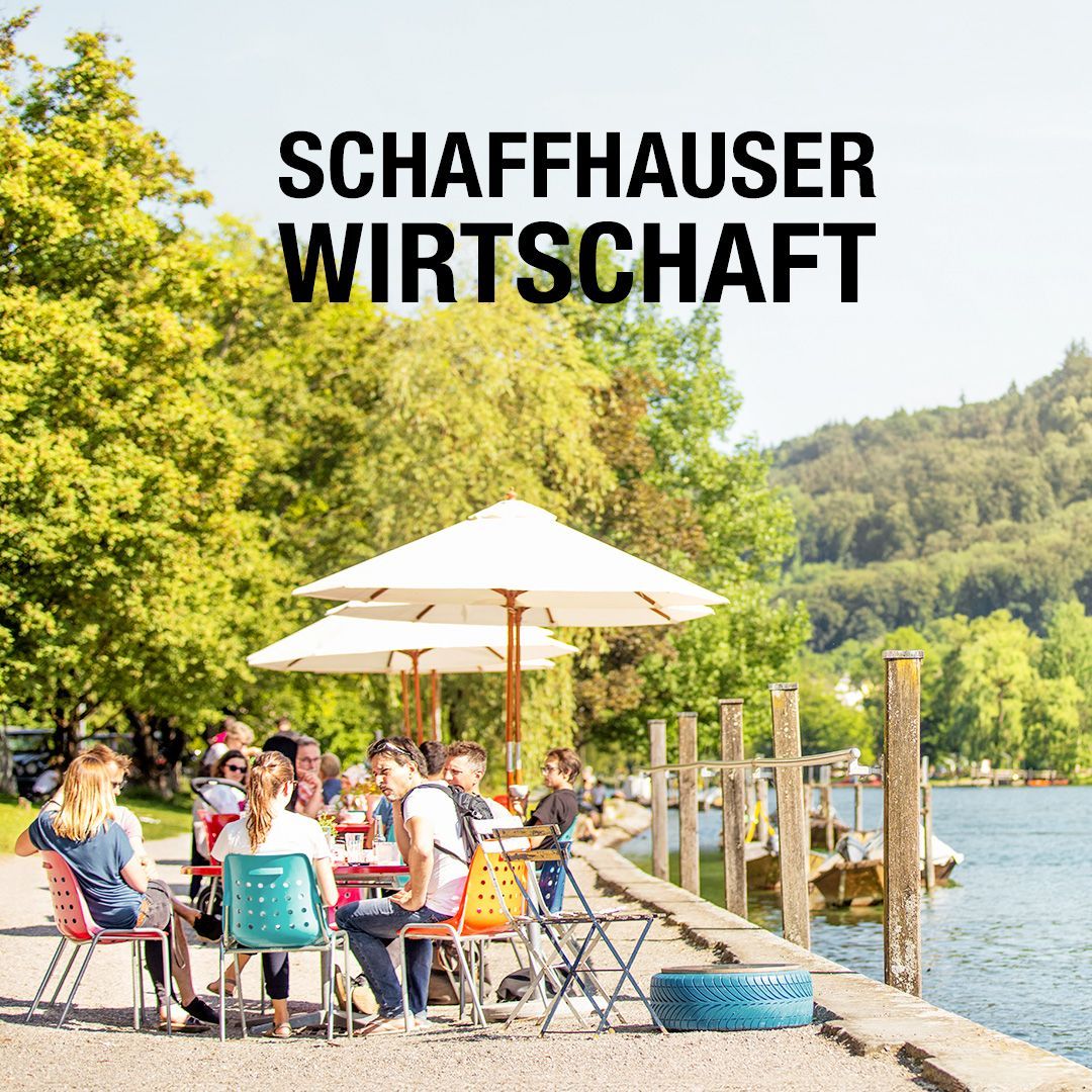 Eine Gruppe von Menschen sitzt unter Sonnenschirmen an Tischen in der Nähe des Rheins.