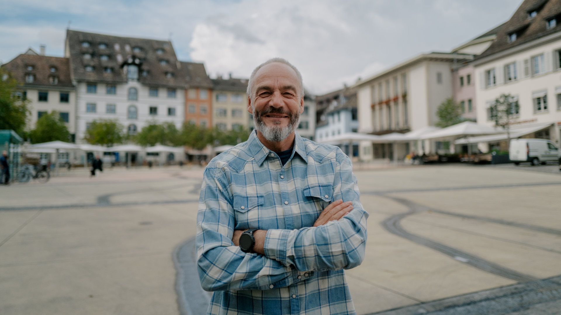 A man with a beard is standing in a city square with his arms crossed.