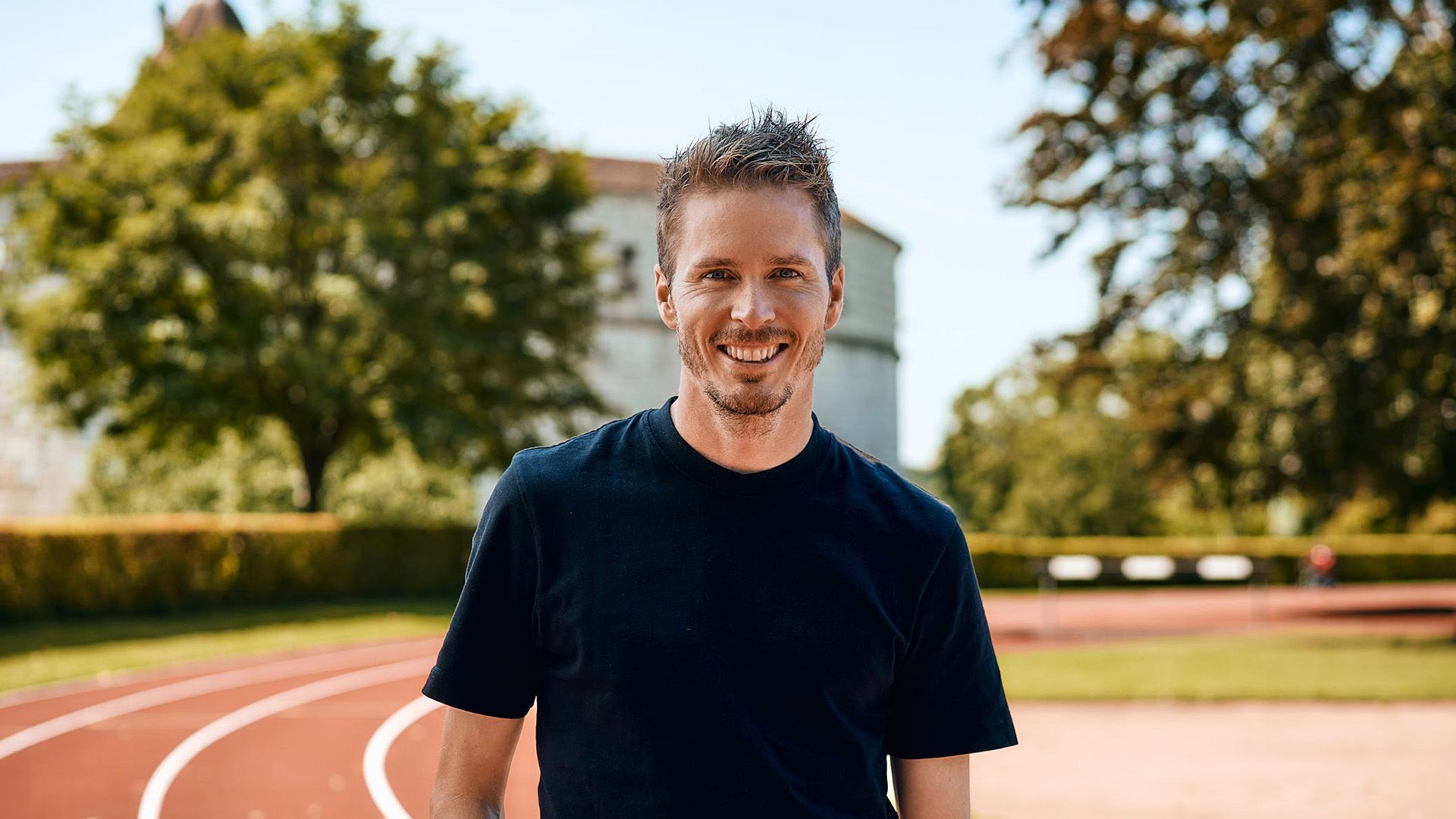 A man in a black shirt is standing on a track and smiling.