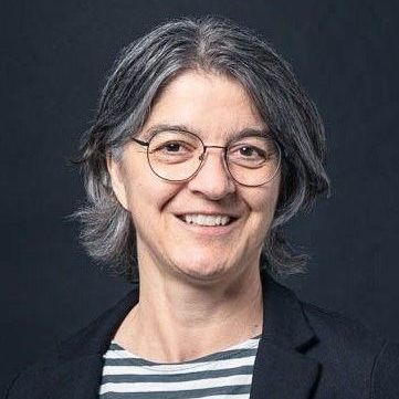 Trix Brunner, with short gray hair wearing glasses and a striped shirt, smiling at the camera against a dark background.